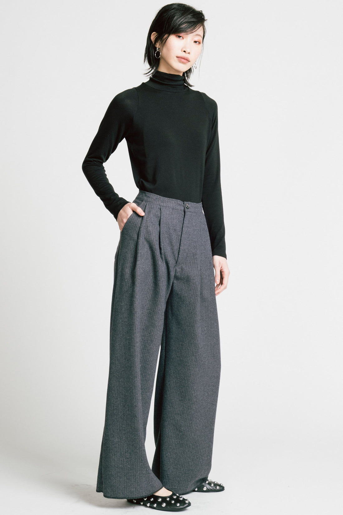 A woman wearing the Aches Turtleneck by Allison Wonderland in Black, a slim fitting classic turtleneck. She is wearing it with the Memory Pants in Denim Pinstripe and standing in front of a white background. 