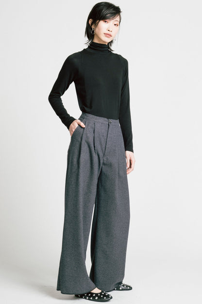 A woman wearing the Aches Turtleneck by Allison Wonderland in Black, a slim fitting classic turtleneck. She is wearing it with the Memory Pants in Denim Pinstripe and standing in front of a white background. 