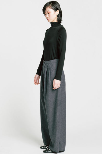 A woman wearing the Aches Turtleneck by Allison Wonderland in Black, a slim fitting classic turtleneck. She is wearing it with the Memory Pants in Denim Pinstripe and standing in front of a white background. 