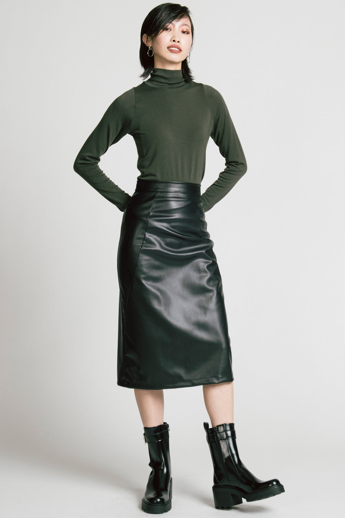 A woman wearing the Aches Turtleneck by Allison Wonderland in Green, a slim fitting classic turtleneck. She is wearing it with the Desire Skirt in Black and standing in front of a white background. 