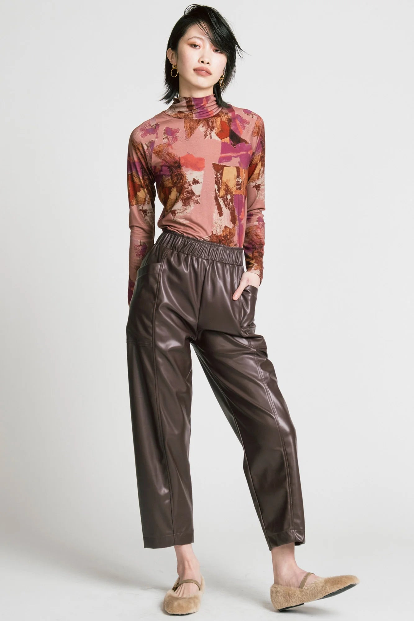A woman wearing the Aches Turtleneck by Allison Wonderland in Burgundy print, a slim fitting classic turtleneck in an abstract pattern. She is wearing it with brown faux leather pants and standing in front of a white background. 