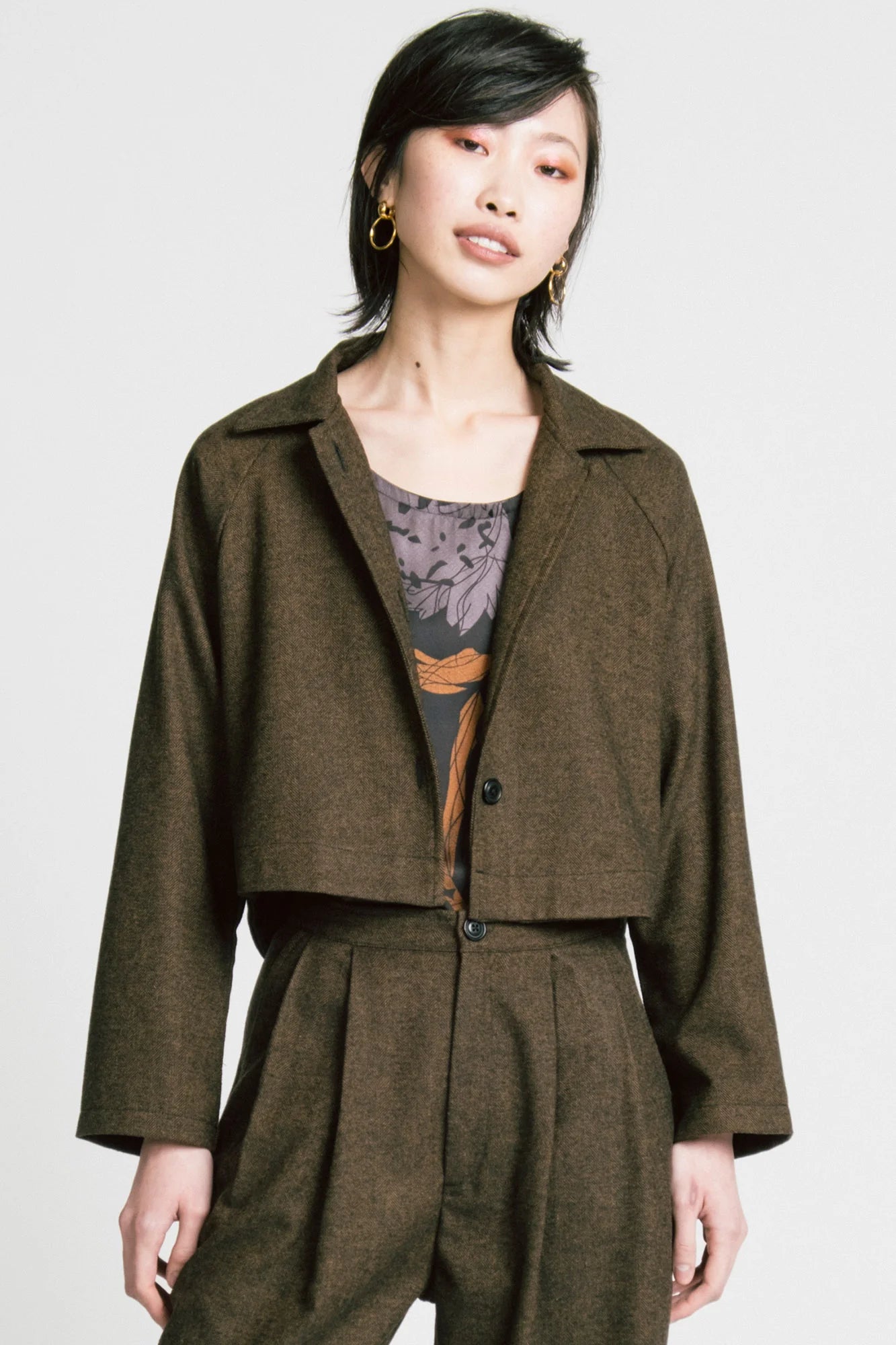 3/4 view of a woman wearing the Cycle Jacket by Allison Wonderland in brown Herringbone, a cropped jacket with a lapel collar, button front, and raglan sleeves. She is wearing it with the Flash top in Horse print and the matching Memory Pants and standing in front of a white background. 