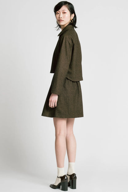 Side view of a woman wearing the Cycle Jacket by Allison Wonderland in brown Herringbone, a cropped jacket with a lapel collar, button front, and raglan sleeves. She is wearing it with a matching skirt and standing in front of a white background. 