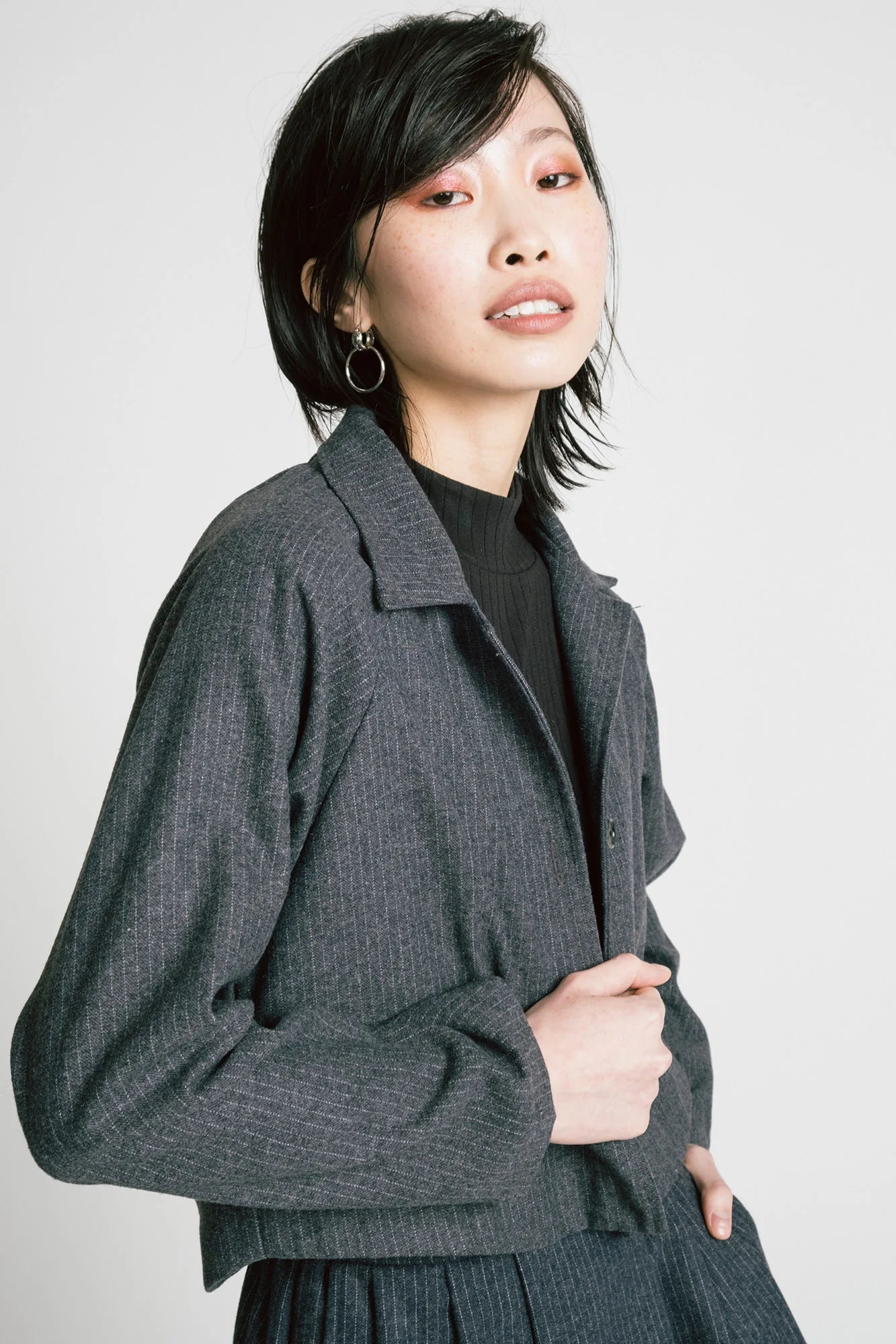 Waist up view of a woman wearing the Cycle Jacket by Allison Wonderland in denim Pinstripe, a cropped jacket with a lapel collar, button front, and raglan sleeves. She is wearing it with a matching skirt and standing in front of a white background. 