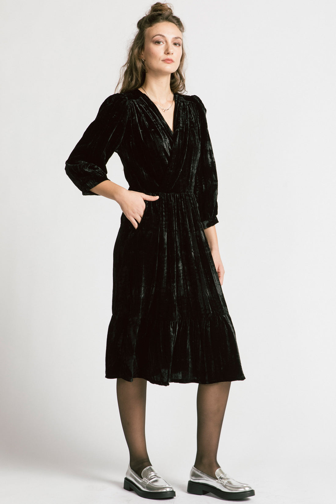 A woman wearing the Dark Lover Dress by Allison Wonderland in Black crushed velvet, featuring a faux-wrap front, 3/4 gathered sleeves, and a tiered midi-length skirt. She is standing in front of a white background. 