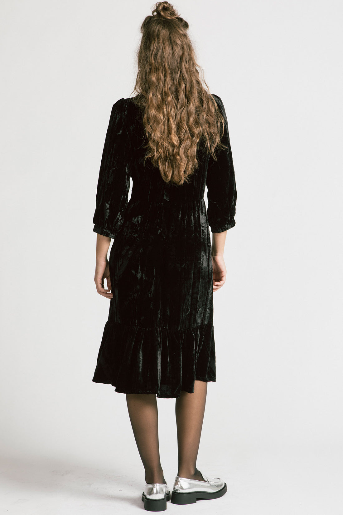 Back view of a woman wearing the Dark Lover Dress by Allison Wonderland in Black crushed velvet, featuring a faux-wrap front, 3/4 gathered sleeves, and a tiered midi-length skirt. She is standing in front of a white background. 