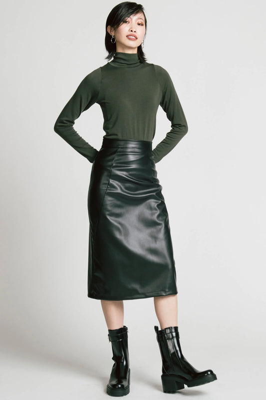 A woman wearing the Desire Skirt by Allison Wonderland in Black faux leather, a midi-length pencil skirt with a two-way back zipper. She is wearing it with a green turtleneck and standing in front of a white background. 