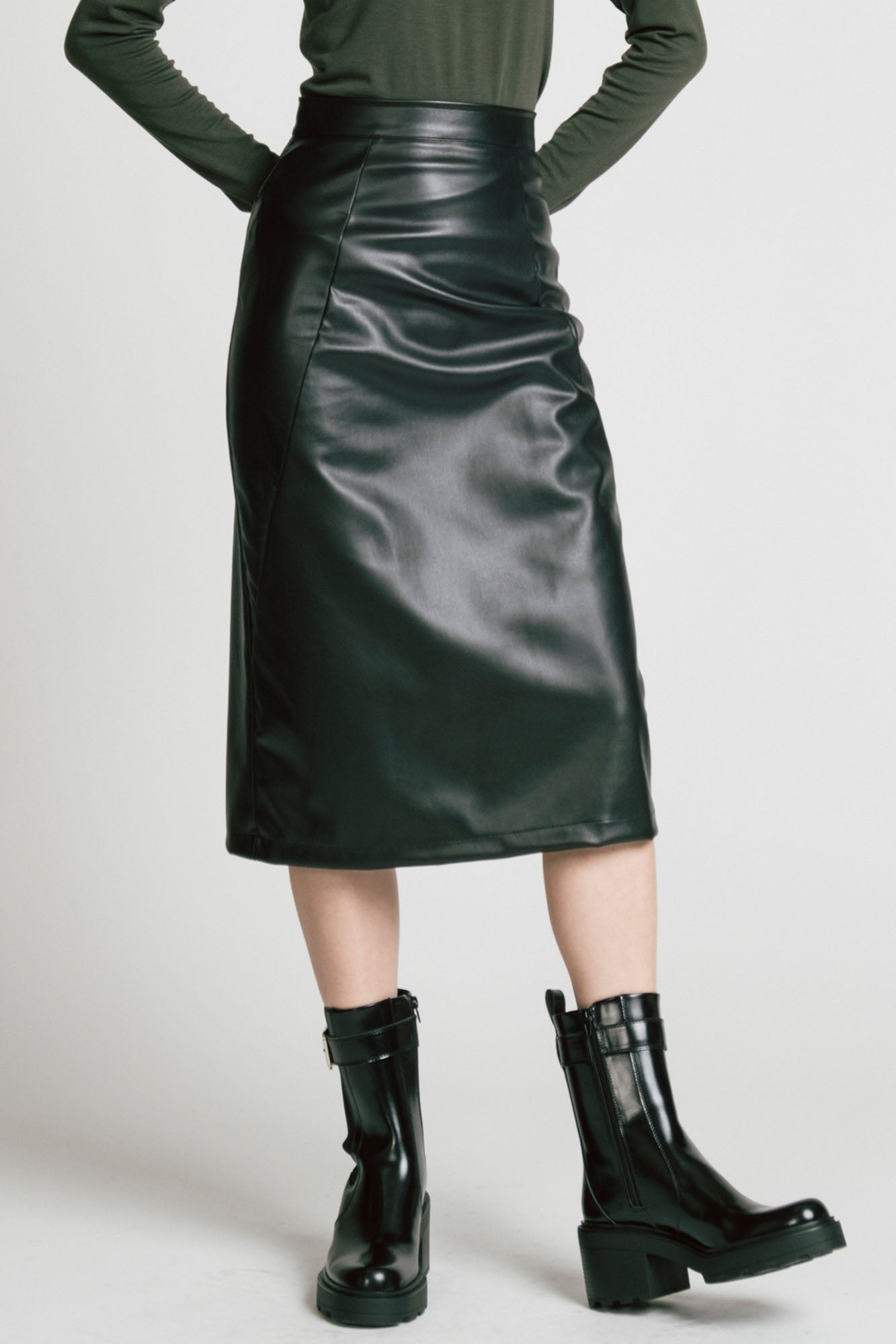 Waist down view of a woman wearing the Desire Skirt by Allison Wonderland in Black faux leather, a midi-length pencil skirt with a two-way back zipper. She is wearing it with a green turtleneck and standing in front of a white background. 