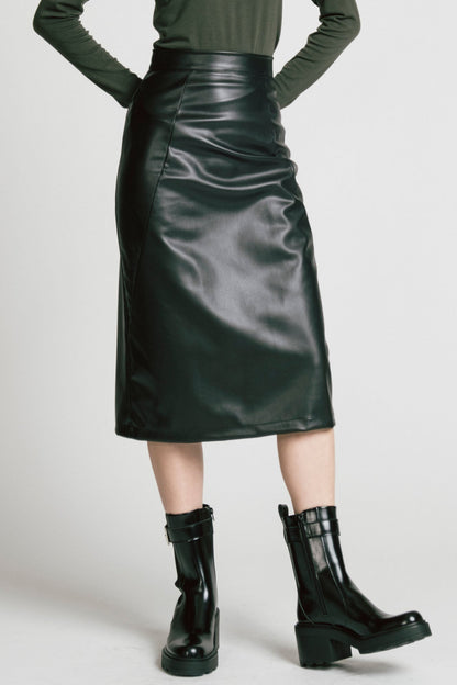 Waist down view of a woman wearing the Desire Skirt by Allison Wonderland in Black faux leather, a midi-length pencil skirt with a two-way back zipper. She is wearing it with a green turtleneck and standing in front of a white background. 