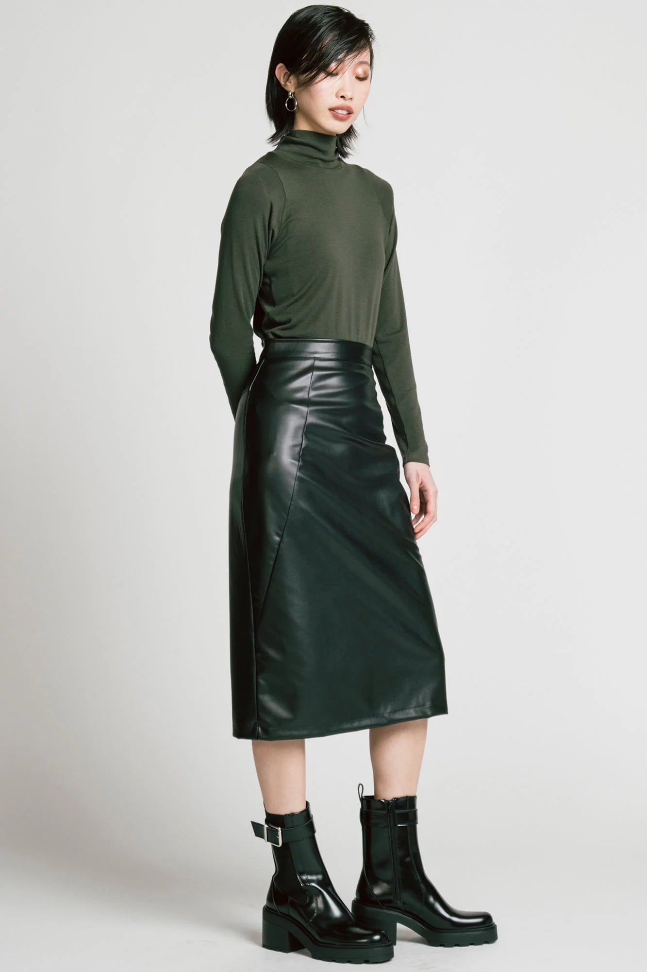 A woman wearing the Desire Skirt by Allison Wonderland in Black faux leather, a midi-length pencil skirt with a two-way back zipper. She is wearing it with a green turtleneck and standing in front of a white background. 