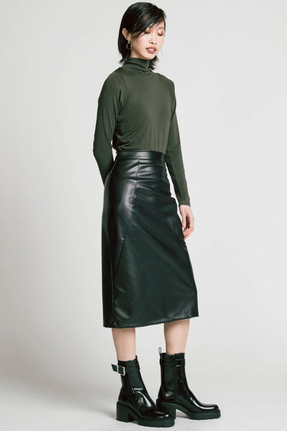 A woman wearing the Desire Skirt by Allison Wonderland in Black faux leather, a midi-length pencil skirt with a two-way back zipper. She is wearing it with a green turtleneck and standing in front of a white background. 