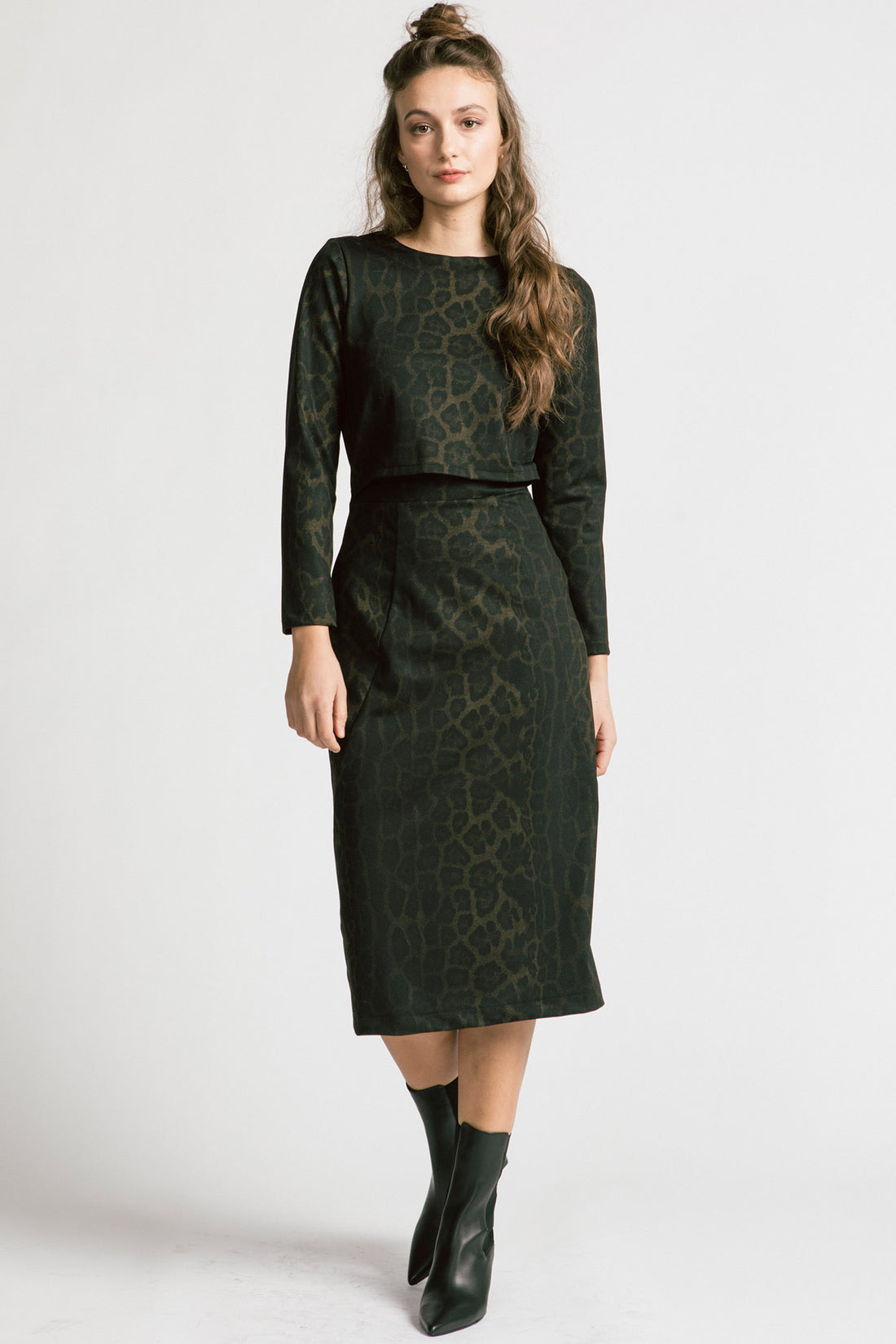  A woman wearing the Allison Wonderland Expectations dress in Leopard, featuring an opening at the front and attached back, a two-way back zipper, V-neckline at the back, long sleeves, and midi-length. She is standing in front of a white background. 