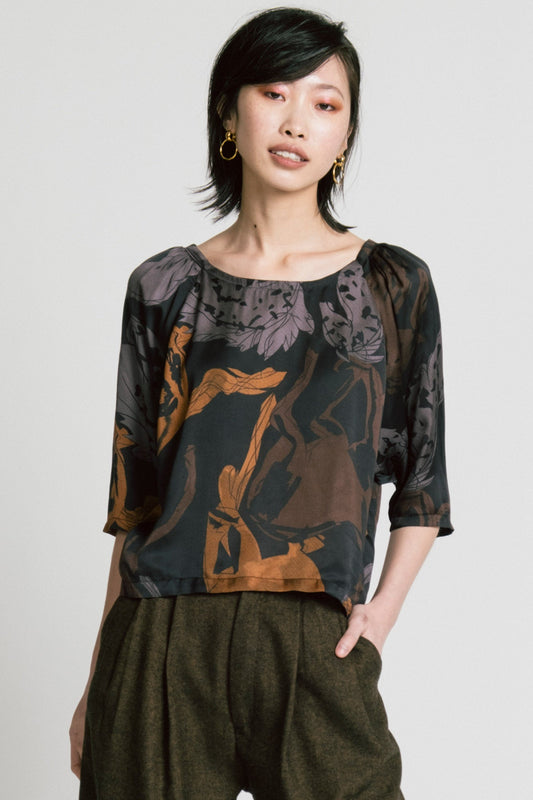3/4 view of a woman wearing the Flash Blouse by Allison Wonderland in Horse Print, featuring 3/4 dropped raglan sleeves, a boat neck with gathers at the shoulders, and a loose fit. She is wearing it with the Memory Pant in Herringbone and standing in front of a white background. 