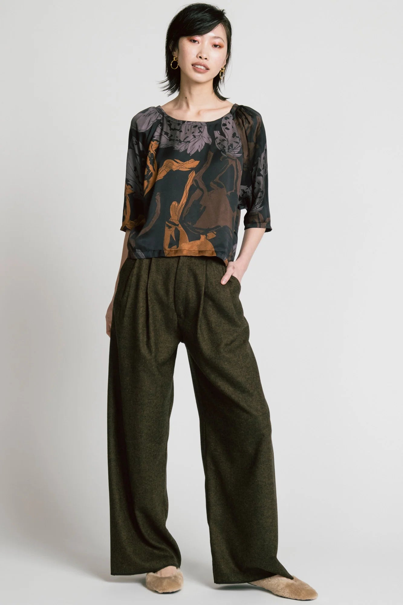 A woman wearing the Flash Blouse by Allison Wonderland in Horse Print, featuring 3/4 dropped raglan sleeves, a boat neck with gathers at the shoulders, and a loose fit. She is wearing it with the Memory Pant in Herringbone and standing in front of a white background. 