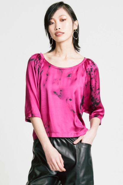 3/4 view of a woman wearing the Flash Blouse by Allison Wonderland in Pink Print, featuring 3/4 dropped raglan sleeves, a boat neck with gathers at the shoulders, and a loose fit. She is wearing it with black faux leather pants and standing in front of a white background.