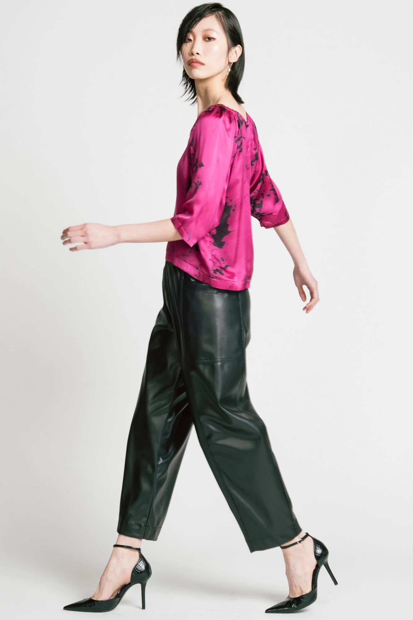 Side view of a woman wearing the Flash Blouse by Allison Wonderland in Pink Print, featuring 3/4 dropped raglan sleeves, a boat neck with gathers at the shoulders, and a loose fit. She is wearing it with black faux leather pants and standing in front of a white background.