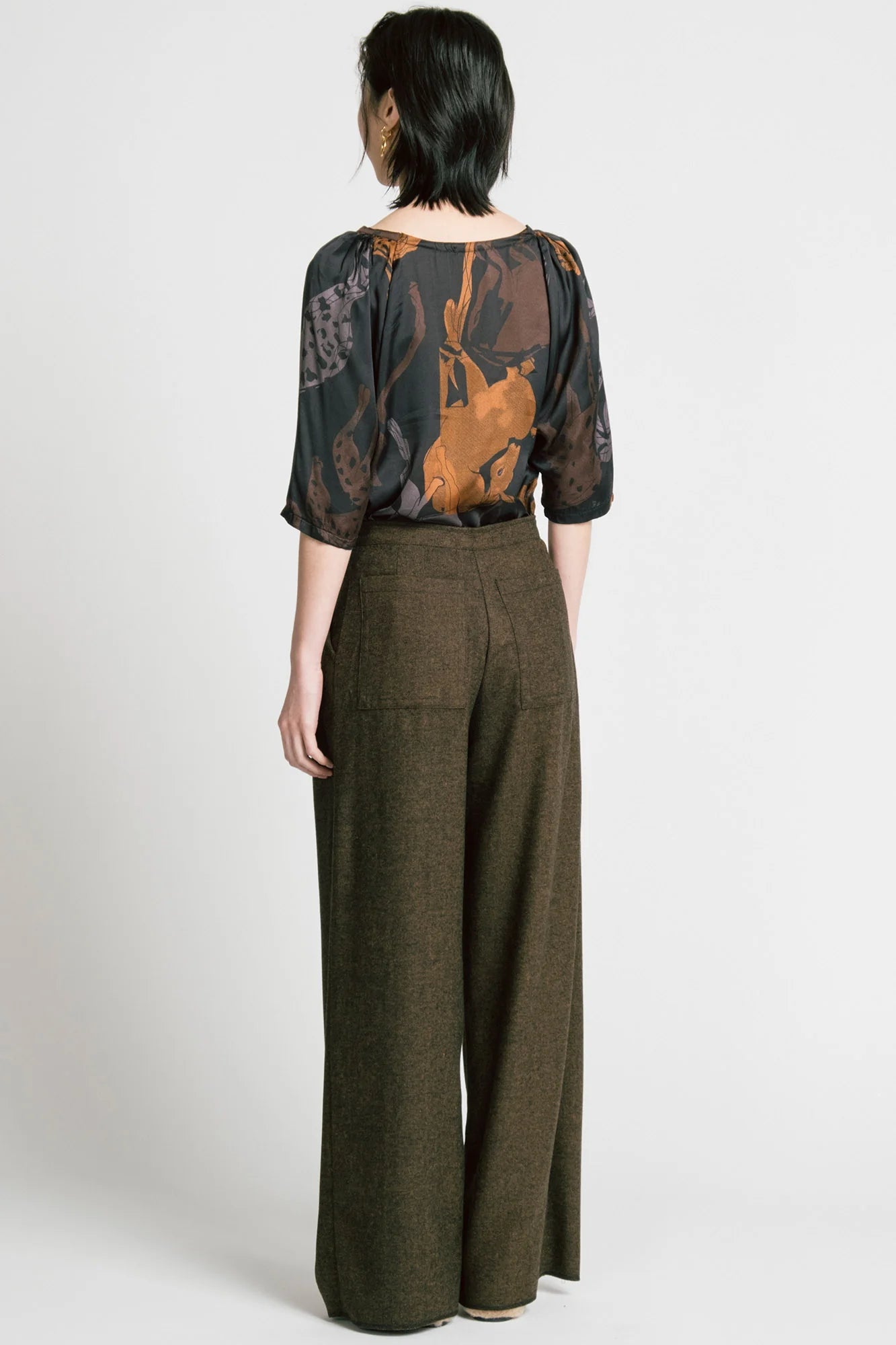 Back view of a woman wearing the Memory Pant by Allison Wonderland in brown Herringbone print, wide-legged pants with front pleats, a zip and button closure, front pockets and back patch pockets. She is wearing them with the Flash Blouse in Horses print and standing in front of a white background. 