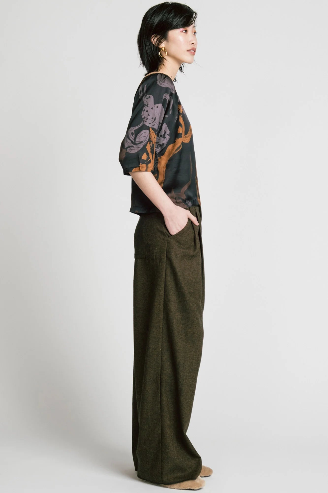 Side view of a woman wearing the Memory Pant by Allison Wonderland in brown Herringbone print, wide-legged pants with front pleats, a zip and button closure, front pockets and back patch pockets. She is wearing them with the Flash Blouse in Horses print and standing in front of a white background. 