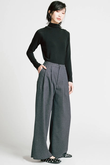 A woman wearing the Memory Pant by Allison Wonderland in denim Pinstripe print, wide-legged pants with front pleats, a zip and button closure, front pockets and back patch pockets. She is wearing them with a black turtleneck and standing in front of a white background. 