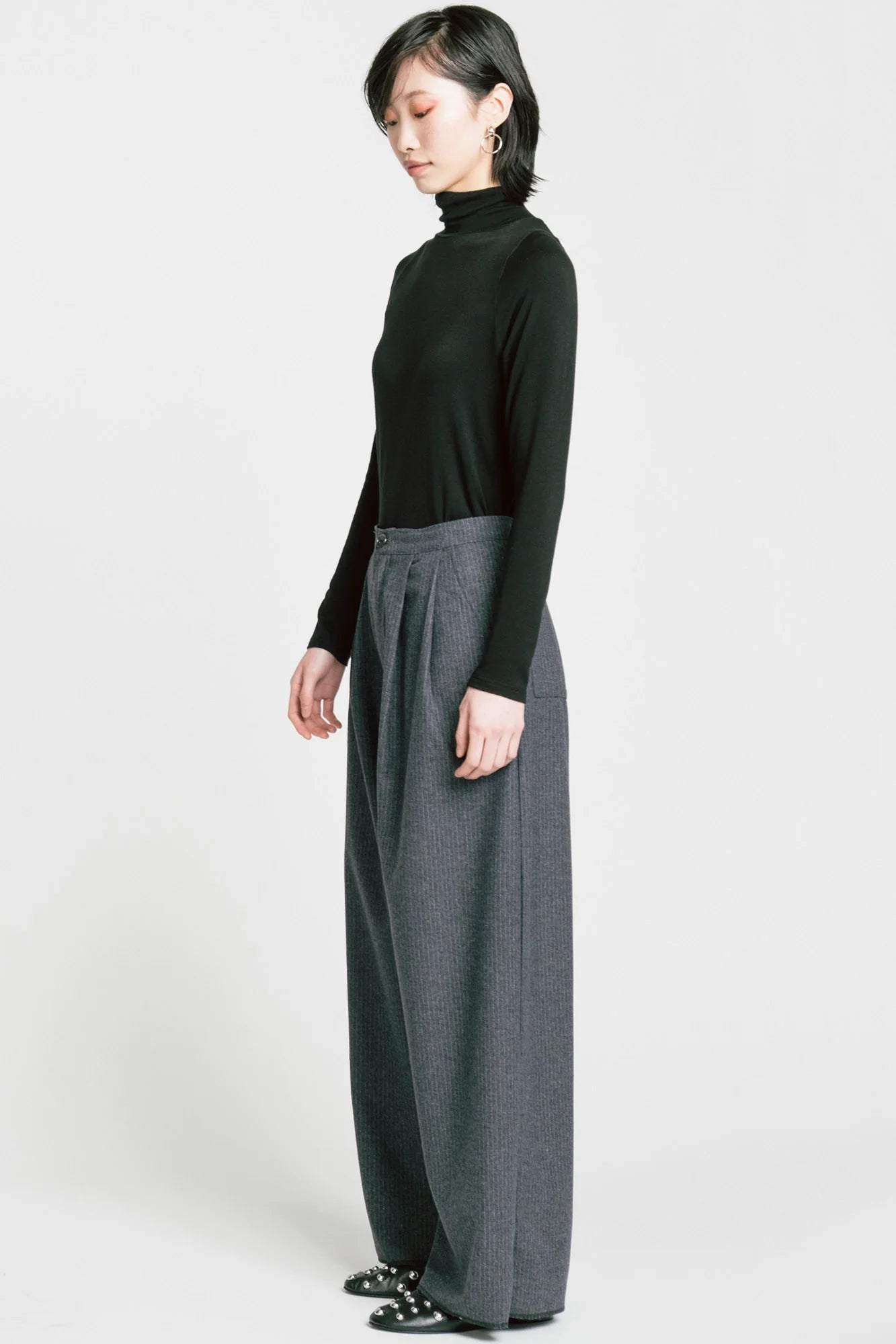 Side view of a woman wearing the Memory Pant by Allison Wonderland in denim Pinstripe print, wide-legged pants with front pleats, a zip and button closure, front pockets and back patch pockets. She is wearing them with a black turtleneck and standing in front of a white background. 