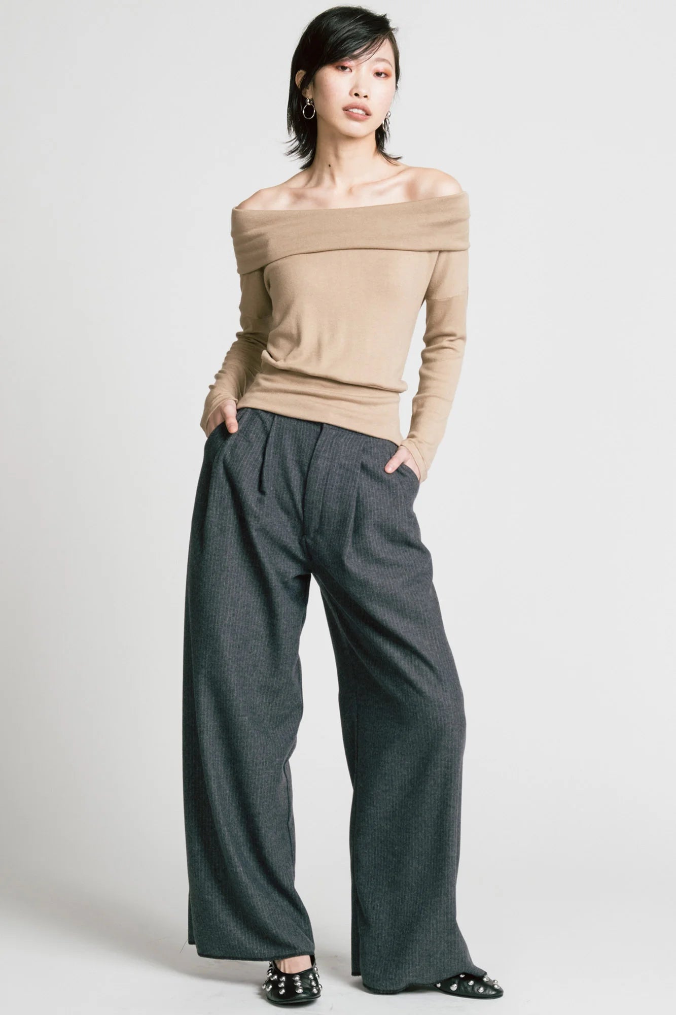 A woman wearing the Memory Pant by Allison Wonderland in denim Pinstripe print, wide-legged pants with front pleats, a zip and button closure, front pockets and back patch pockets. She is wearing them with a beige top and standing in front of a white background. 