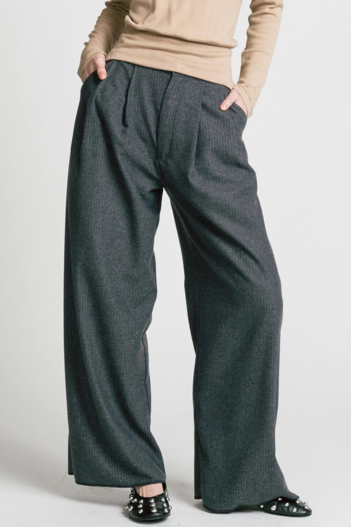 Waist-down view of a woman wearing the Memory Pant by Allison Wonderland in denim Pinstripe print, wide-legged pants with front pleats, a zip and button closure, front pockets and back patch pockets. She is wearing them with a beige top and standing in front of a white background. 