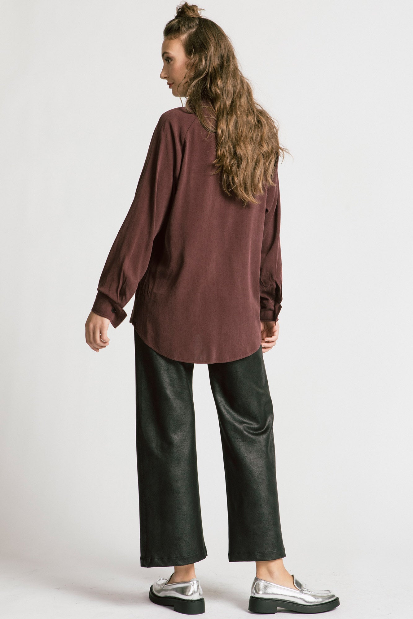 Back view of a woman wearing the Shanna Pants by Allison Wonderland in Black Leatherette, loose fitting flared pants with an elastic waistband. She is wearing them with a brown top and standing in front of a silver background. 