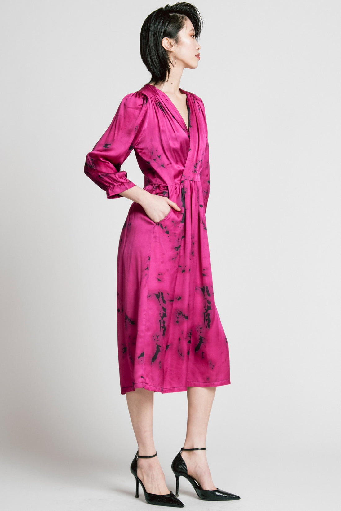 Side view of a woman wearing the Therapy Dress by Allison Wonderland in Pink Print, with a faux wrap front, gathers at the shoulders cuffs and waist, and a full midi-length skirt. She is standing in front of a white background. 