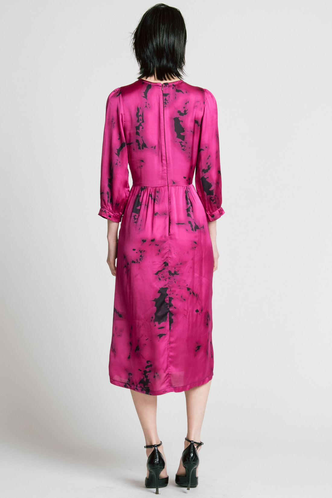 Back view of a woman wearing the Therapy Dress by Allison Wonderland in Pink Print, with a faux wrap front, gathers at the shoulders cuffs and waist, and a full midi-length skirt. She is standing in front of a white background. 