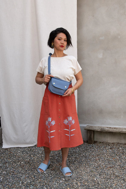  A woman wearing the Nymphea Screenprinted Skirt by Kazak in Red Pysanka with a cream top, she is standing outside in front of white curtain and holding a blue purse.