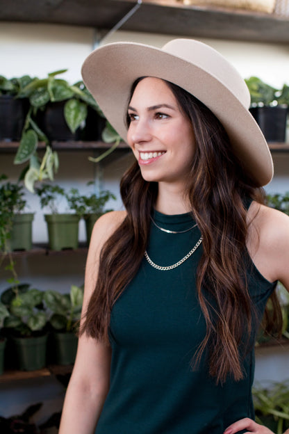 Waist up view of a woman wearing the Bev Tank Dress by Ang Hill in Juniper, a sleeveless, slim-fitting dress in cotton/modal with a high neck and a below the knee hem. She is wearing it with a beige hat and standing in front of shelves of plants. 
