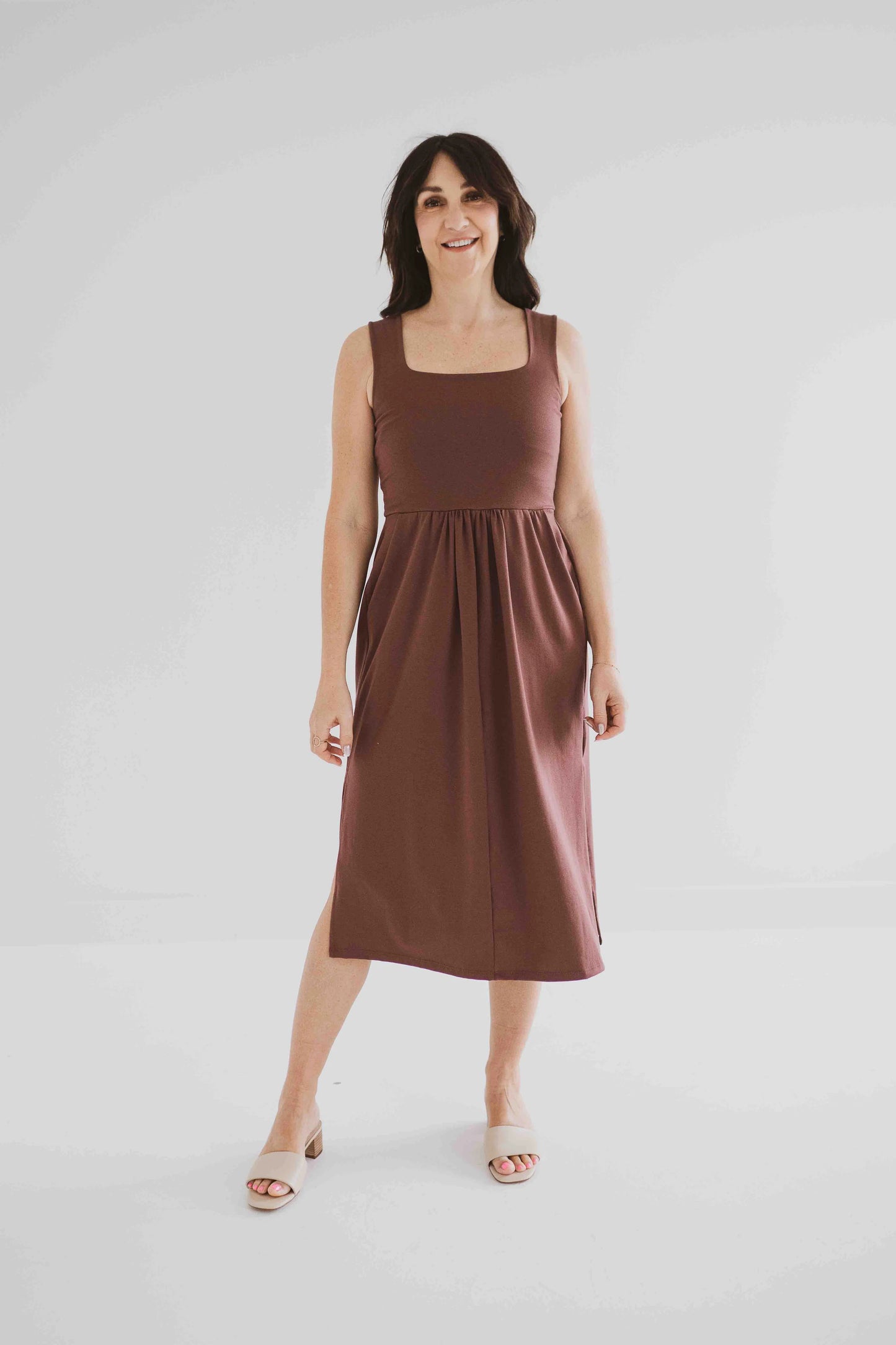 A woman wearing the Audrey Tank Dress by Ang Hill in Mocha, featuring a square neckline, wide straps, a fitted bodice, a skirt that gathers at the waist and falls below the knee with two side slits, and pockets. She is standing in front of a white background. 
