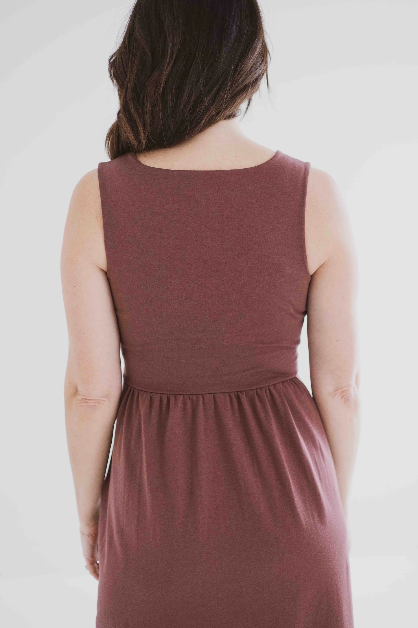 Back 3/4 view of a woman wearing the Audrey Tank Dress by Ang Hill in Mocha, featuring a square neckline, wide straps, a fitted bodice, a skirt that gathers at the waist and falls below the knee with two side slits, and pockets. She is standing in front of a white background. 