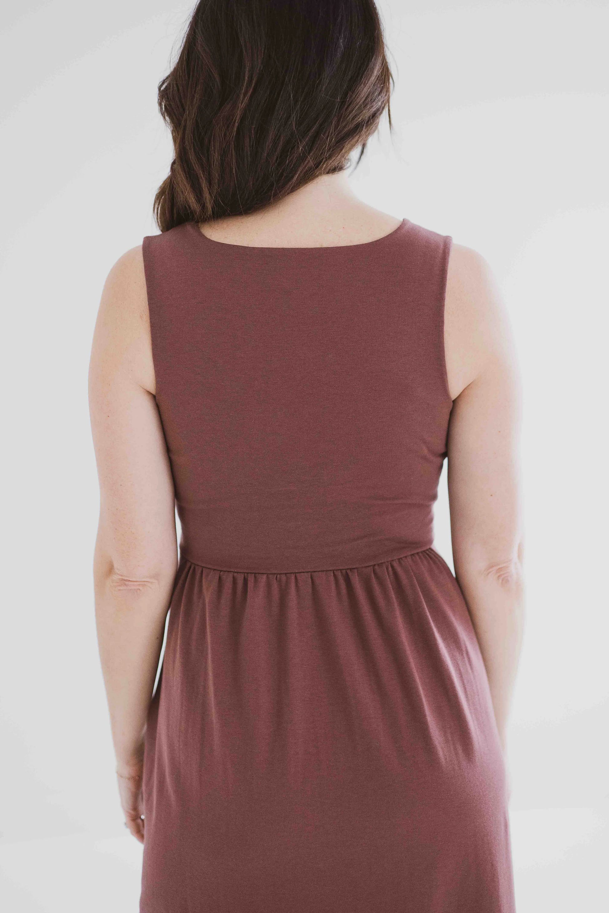 Back 3/4 view of a woman wearing the Audrey Tank Dress by Ang Hill in Mocha, featuring a square neckline, wide straps, a fitted bodice, a skirt that gathers at the waist and falls below the knee with two side slits, and pockets. She is standing in front of a white background. 