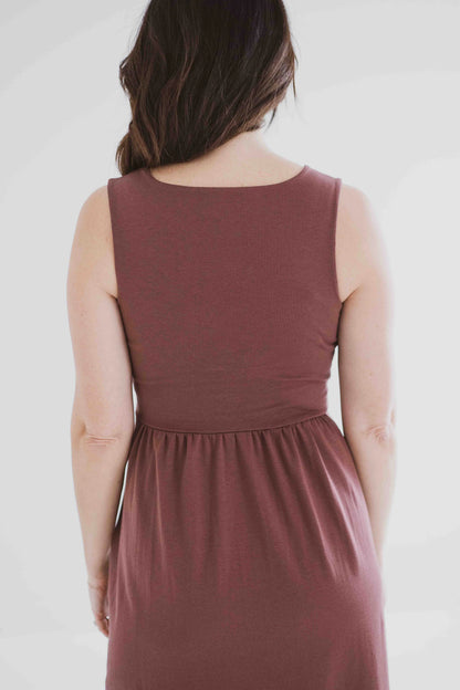 Back 3/4 view of a woman wearing the Audrey Tank Dress by Ang Hill in Mocha, featuring a square neckline, wide straps, a fitted bodice, a skirt that gathers at the waist and falls below the knee with two side slits, and pockets. She is standing in front of a white background. 