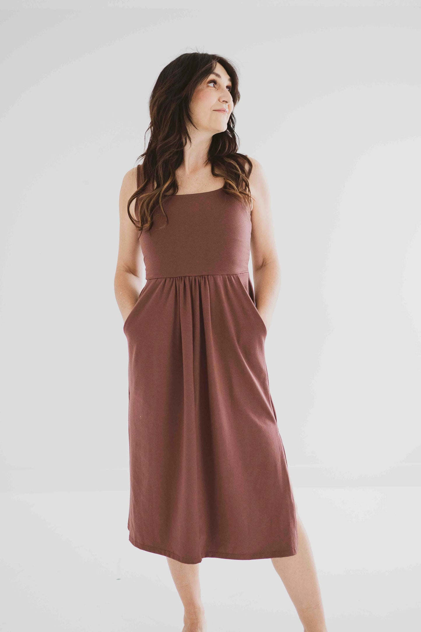 A woman wearing the Audrey Tank Dress by Ang Hill in Mocha, featuring a square neckline, wide straps, a fitted bodice, a skirt that gathers at the waist and falls below the knee with two side slits, and pockets. She is standing in front of a white background. 
