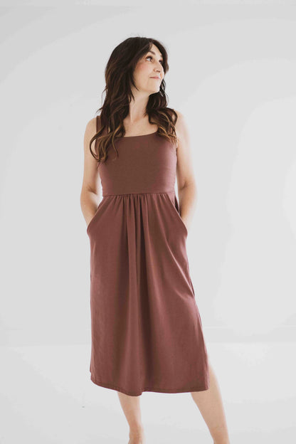 A woman wearing the Audrey Tank Dress by Ang Hill in Mocha, featuring a square neckline, wide straps, a fitted bodice, a skirt that gathers at the waist and falls below the knee with two side slits, and pockets. She is standing in front of a white background. 