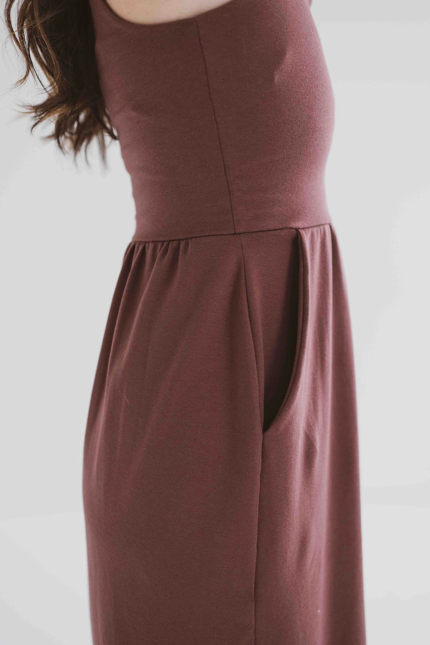 Side 3/4 view of a woman wearing the Audrey Tank Dress by Ang Hill in Mocha, featuring a square neckline, wide straps, a fitted bodice, a skirt that gathers at the waist and falls below the knee with two side slits, and pockets. She is standing in front of a white background. 