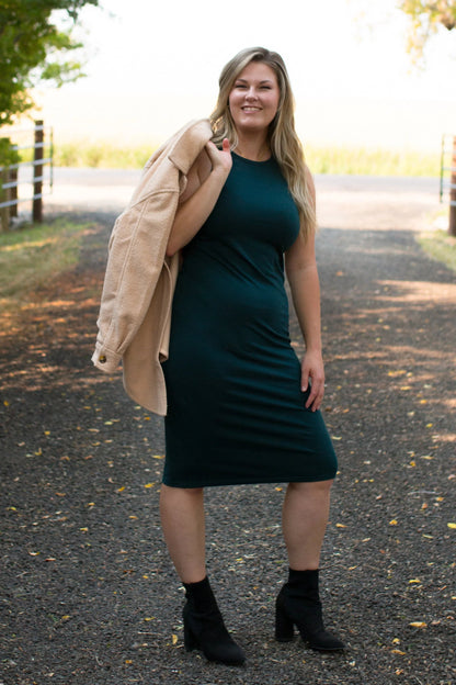 A woman wearing the Bev Tank Dress by Ang Hill in Juniper, a sleeveless, slim-fitting dress in cotton/modal with a high neck and a below the knee hem. She is wearing it with a beige jacket thrown over her shoulder and is standing outside on a gravel path. 