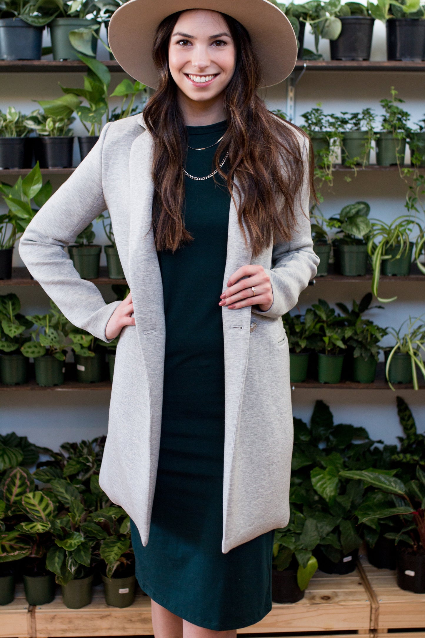 A woman wearing the Bev Tank Dress by Ang Hill in Juniper, a sleeveless, slim-fitting dress in cotton/modal with a high neck and a below the knee hem. She is wearing it with a grey coat and beige hat and standing in front of shelves of plants. 