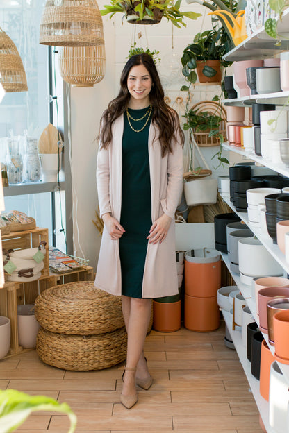 A woman wearing the Bev Tank Dress by Ang Hill in Juniper, a sleeveless, slim-fitting dress in cotton/modal with a high neck and a below the knee hem. She is wearing it with a beige sweater and standing in front of shelves of plants. 