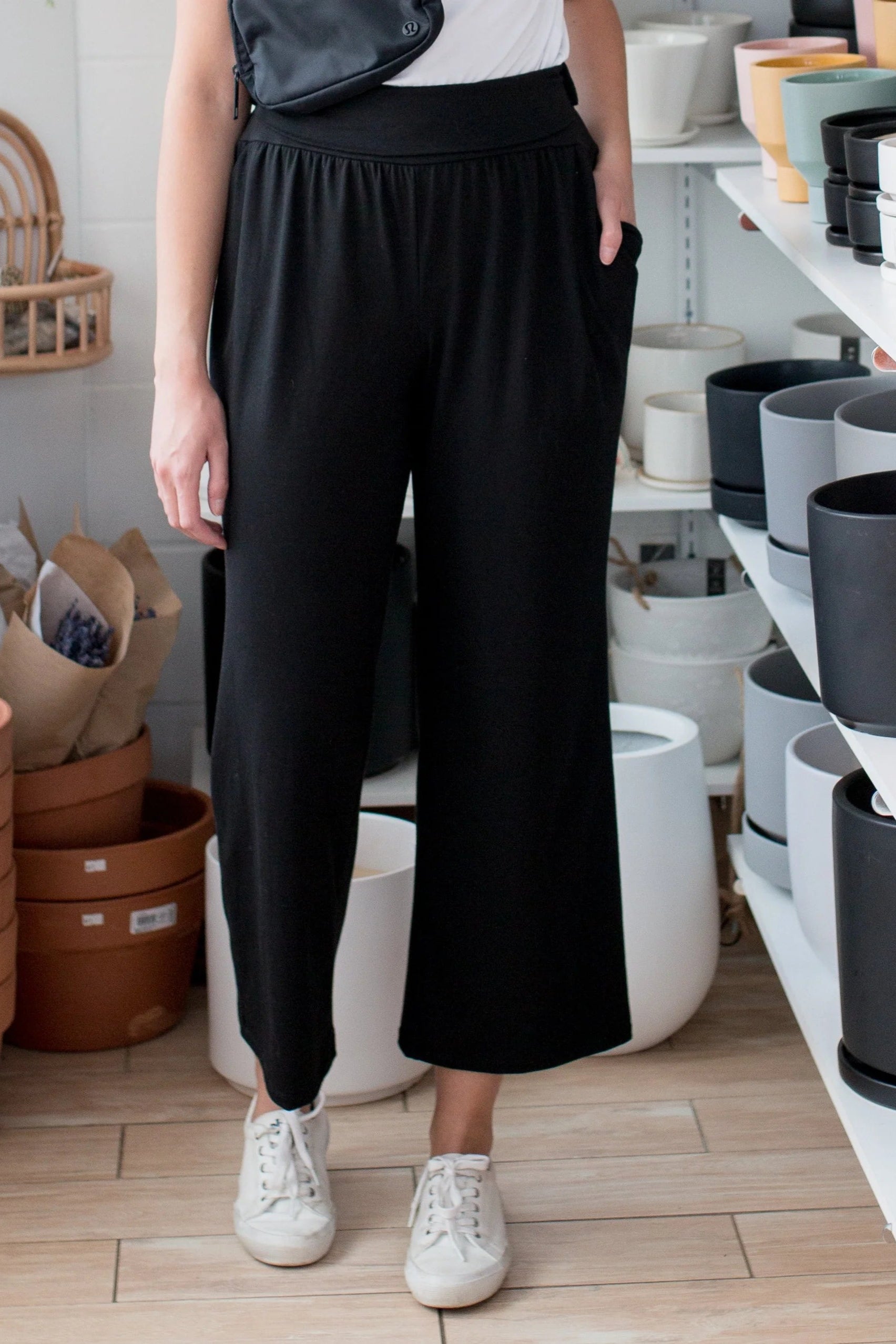 Waist down view of a woman wearing the Bree Wide Leg Pants by Ang Hill in Black, cropped wide-legged pants with a high elastic waist band and deep pockets. She is wearing them with a white t-shirt and standing in a pottery studio. 