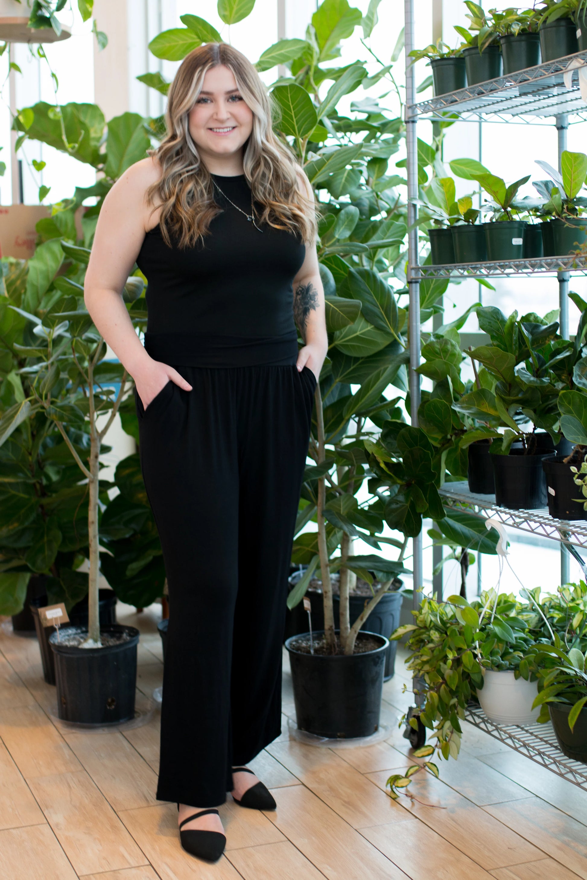 A woman wearing the Bree Wide Leg Pants by Ang Hill in Black, cropped wide-legged pants with a high elastic waist band and deep pockets. She is wearing them with a black tank top and standing in front of plants. 