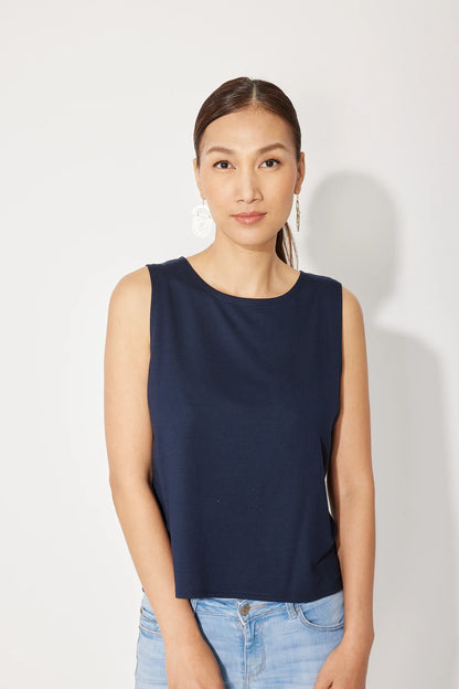 A woman wearing the Baie Cami by Canopée in Navy, a bamboo French Terry tank with a high neck and loose fit. She is wearing it with jeans and standing in front of a white background. 