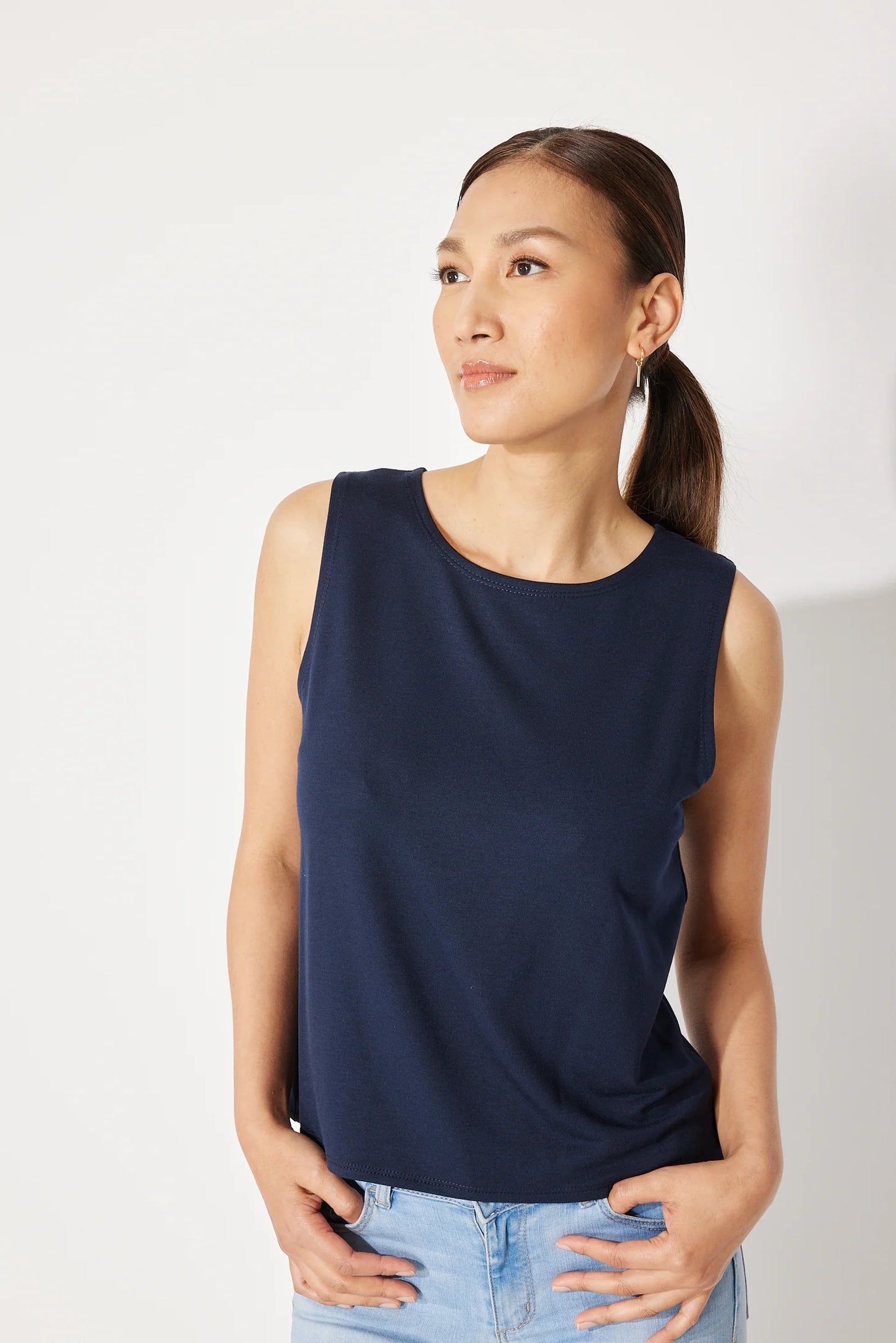 A woman wearing the Baie Cami by Canopée in Navy, a bamboo French Terry tank with a high neck and loose fit. She is wearing it with jeans and standing in front of a white background. 