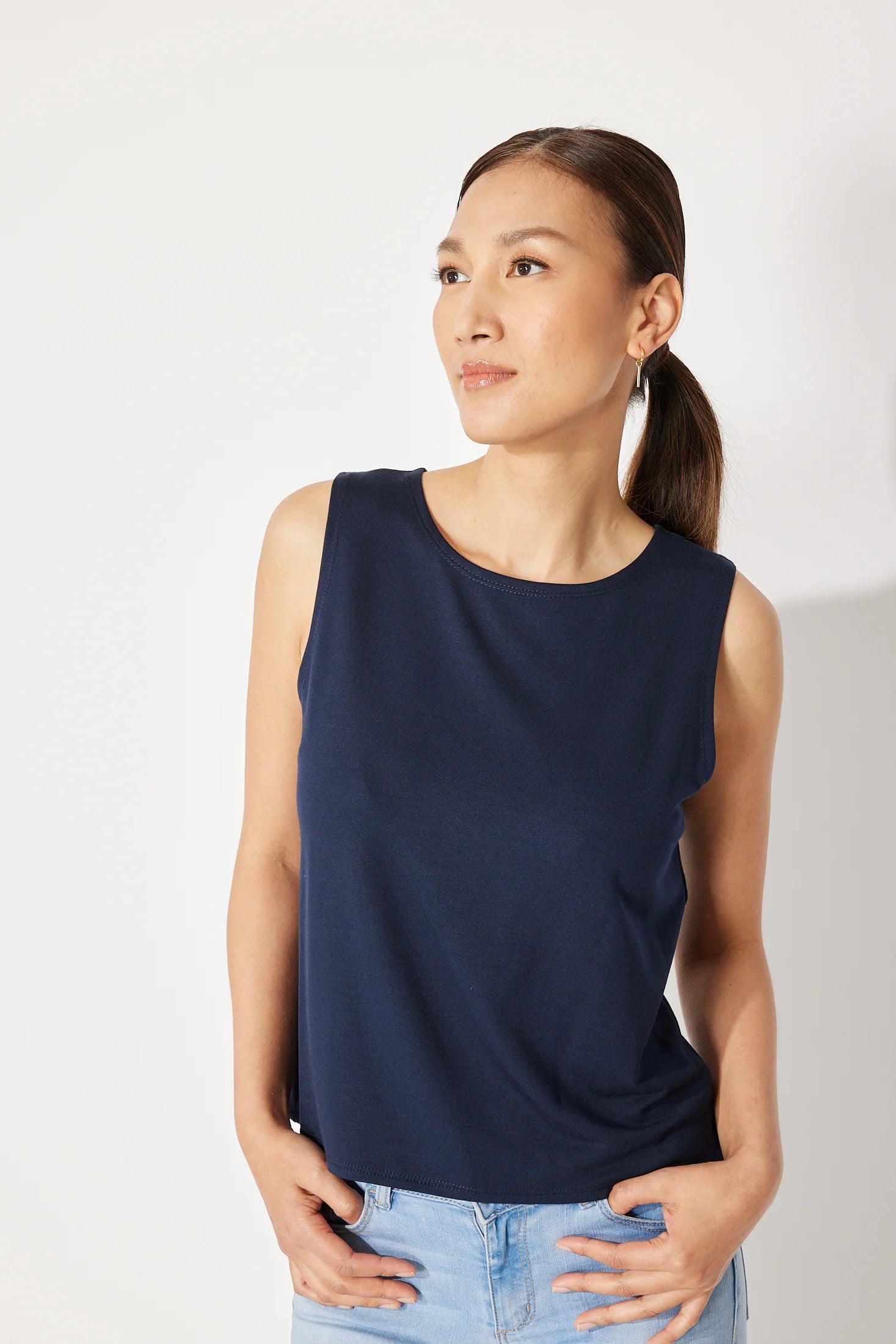 A woman wearing the Baie Cami by Canopée in Navy, a bamboo French Terry tank with a high neck and loose fit. She is wearing it with jeans and standing in front of a white background. 