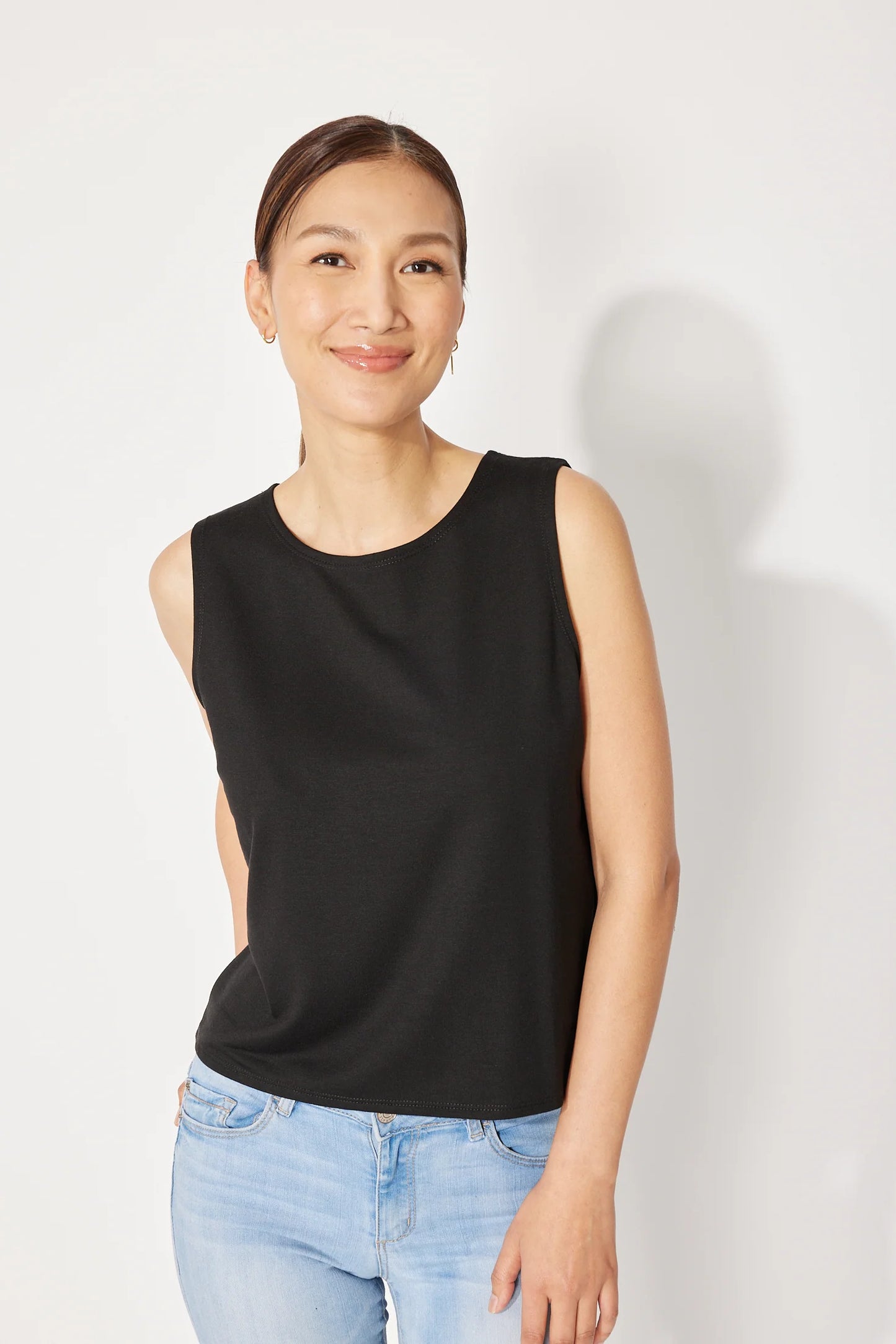 A woman wearing the Baie Cami by Canopée in Black, a bamboo French Terry tank with a high neck and loose fit. She is wearing it with jeans and standing in front of a white background. 