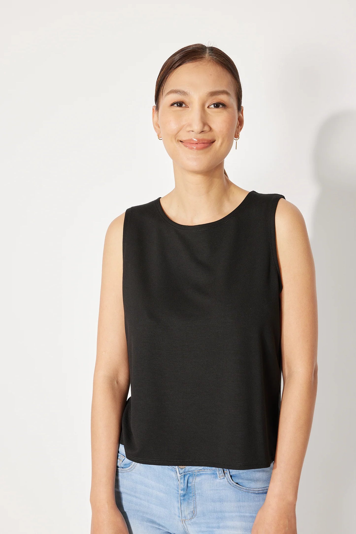 A woman wearing the Baie Cami by Canopée in Black, a bamboo French Terry tank with a high neck and loose fit. She is wearing it with jeans and standing in front of a white background. 