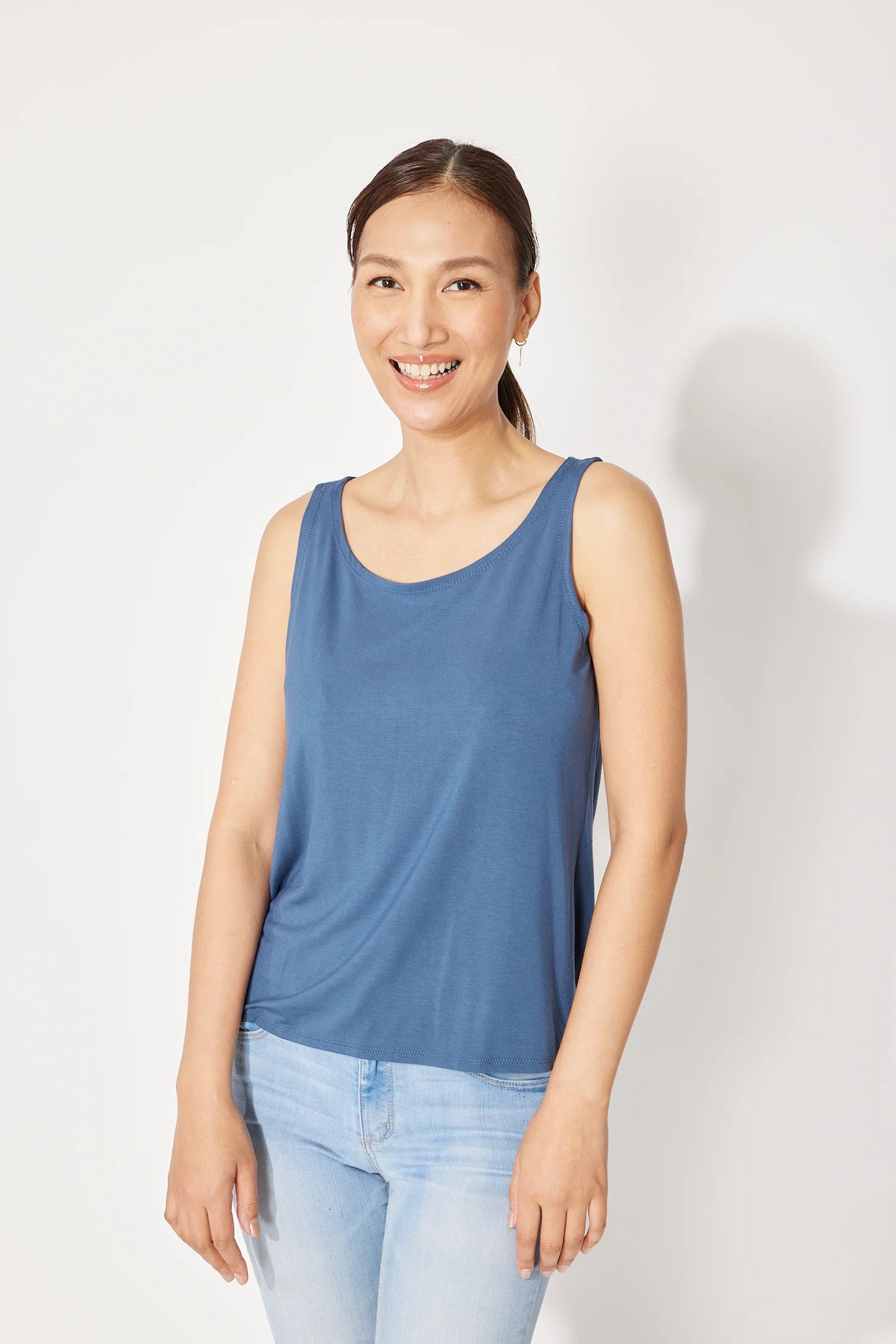 A woman wearing the Saureau Cami by Canopee in Blue, a wide-strapped tank with a scooped neck, loose fit, and hip length. She is wearing it with jeans and standing in front of a white background. 