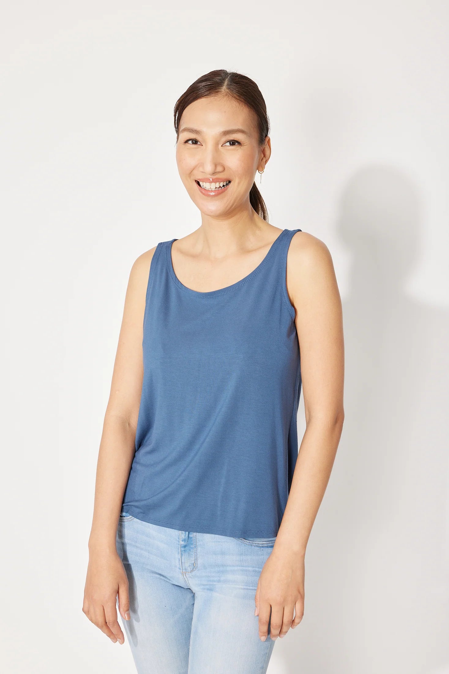 A woman wearing the Saureau Cami by Canopee in Blue, a wide-strapped tank with a scooped neck, loose fit, and hip length. She is wearing it with jeans and standing in front of a white background. 